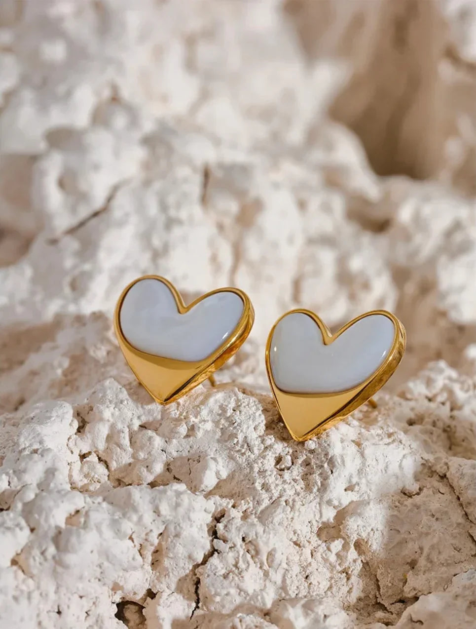 Gold heart-shaped stud earrings with white enamel on textured stone background