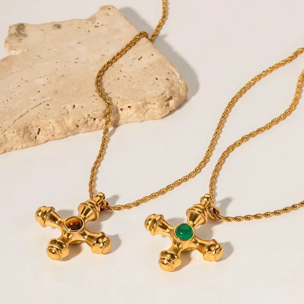 Gold cross necklaces with green and brown gemstones on rope chains, styled on a stone slab.