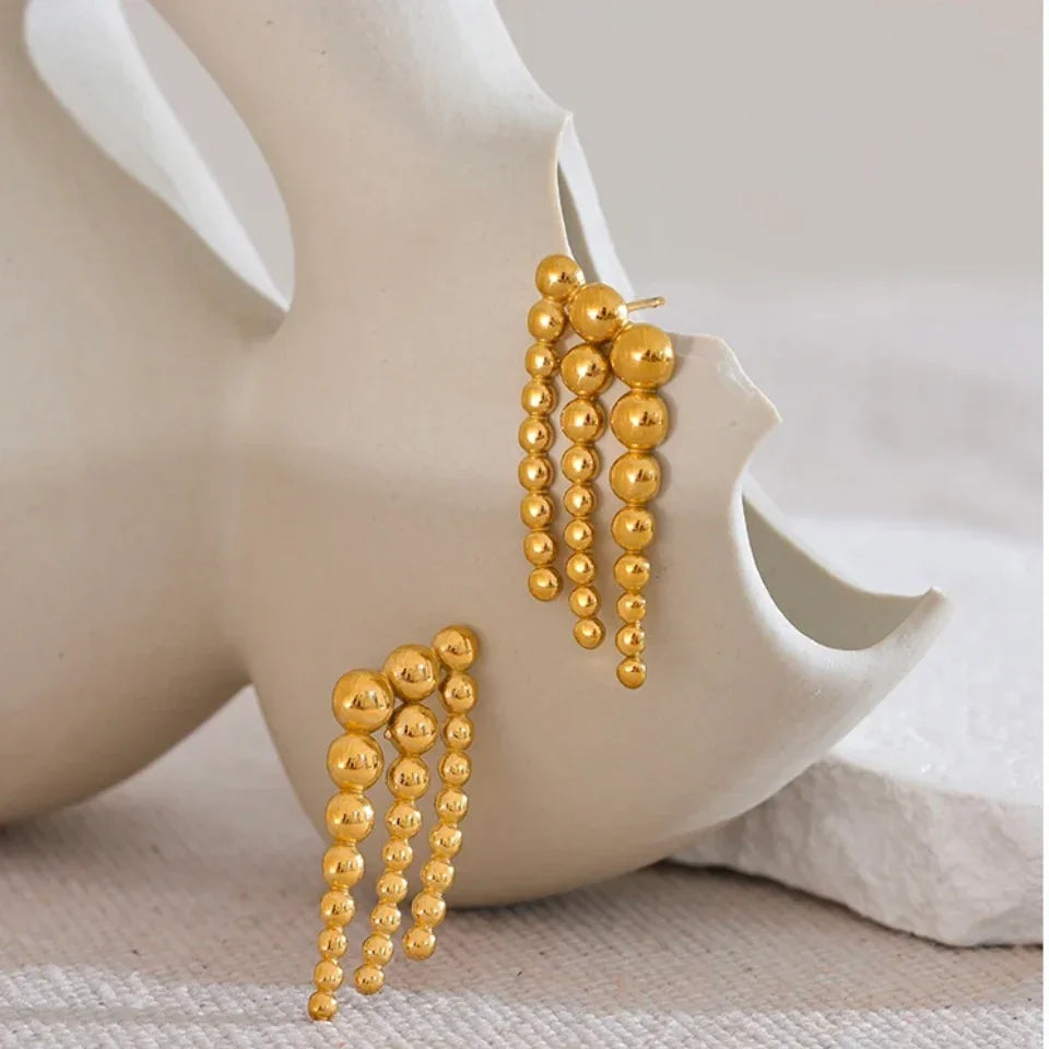Gold statement earrings with cascading round beads displayed on a white ceramic vase.