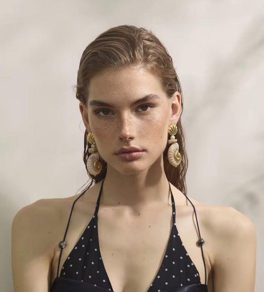 Woman wearing a black polka dot halter bikini and statement shell earrings, neutral background.